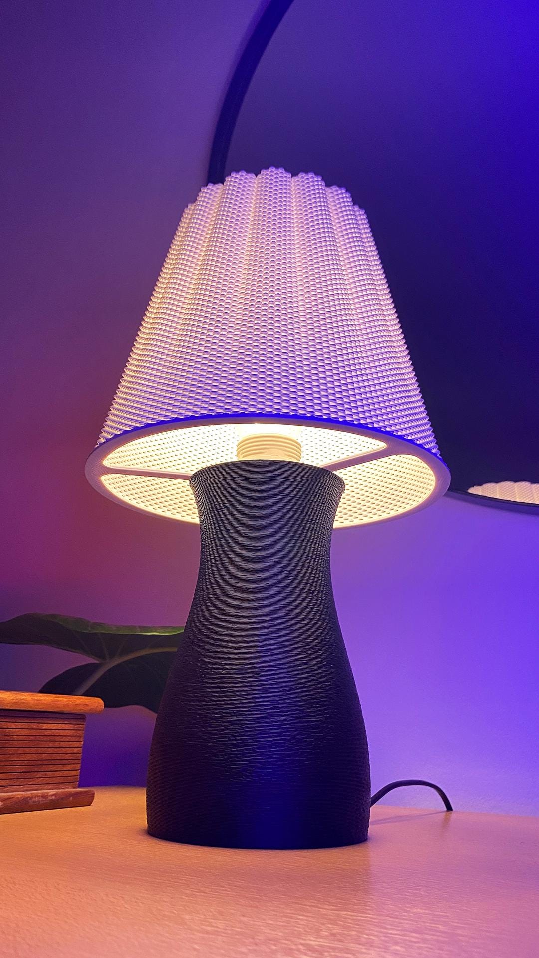 Table lamp with textured black base and white lampshade on a wooden surface with purple background