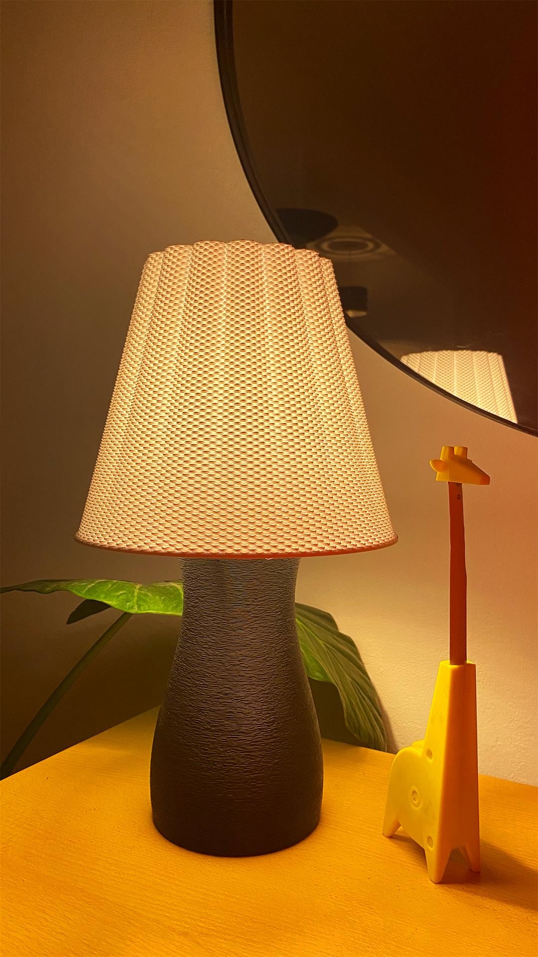 Table lamp with textured white shade on a wooden surface, with a plant and decorative item in the background.