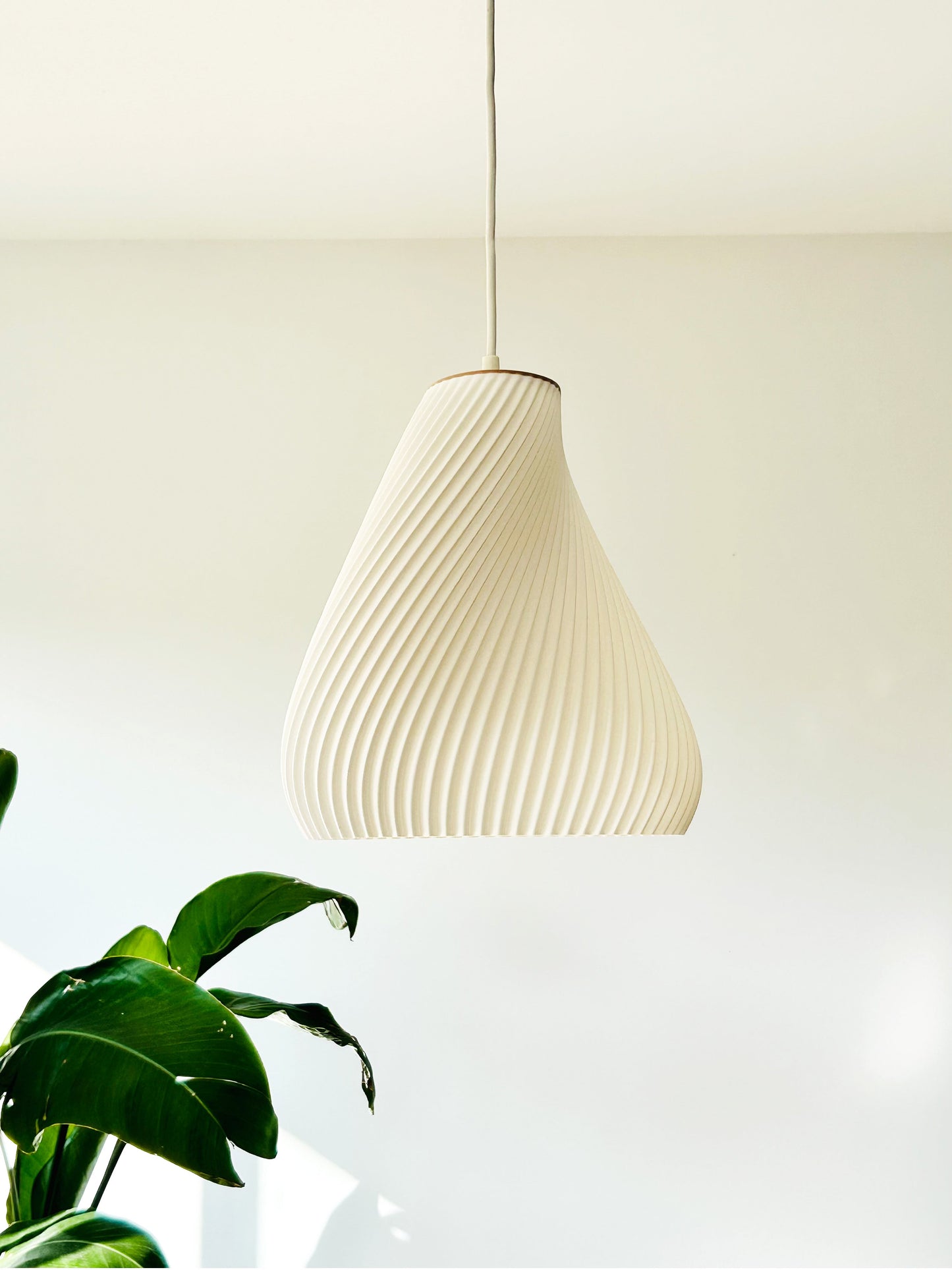White textured pendant light hanging against a white wall with a plant in the foreground.