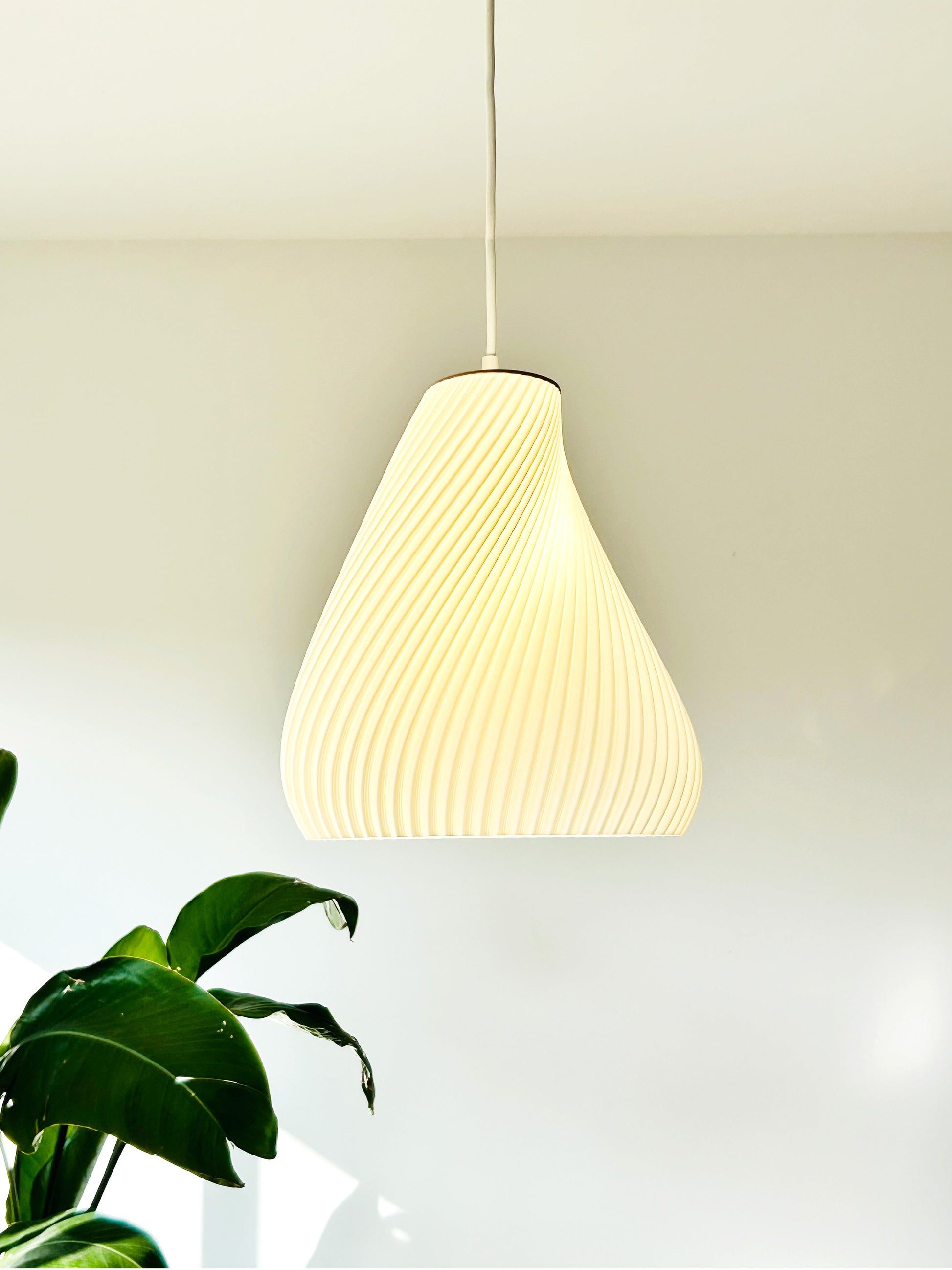 White pendant light hanging against a white wall with a plant in the corner.