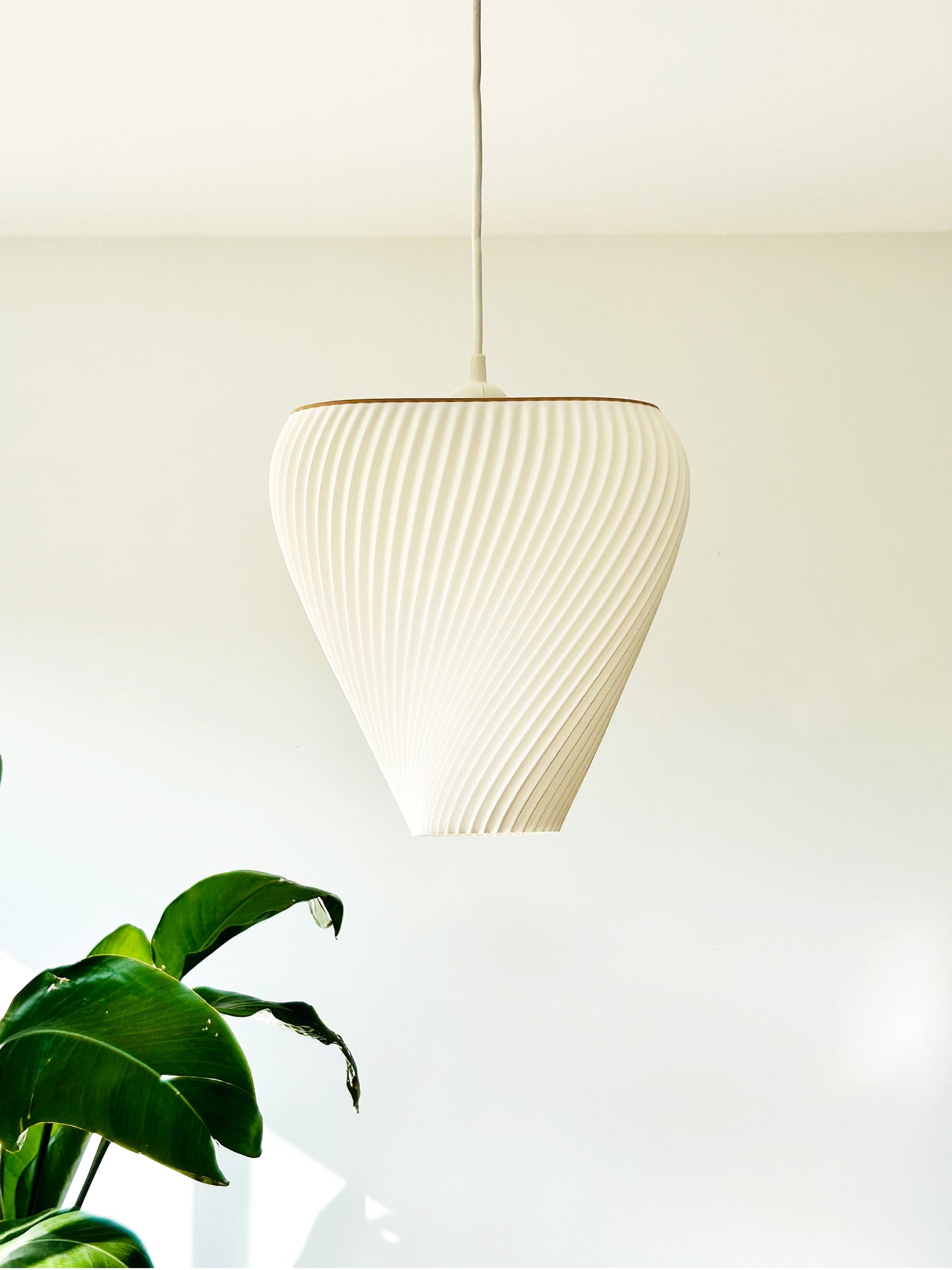 White ribbed pendant light hanging against a plain white wall with a plant in the corner.