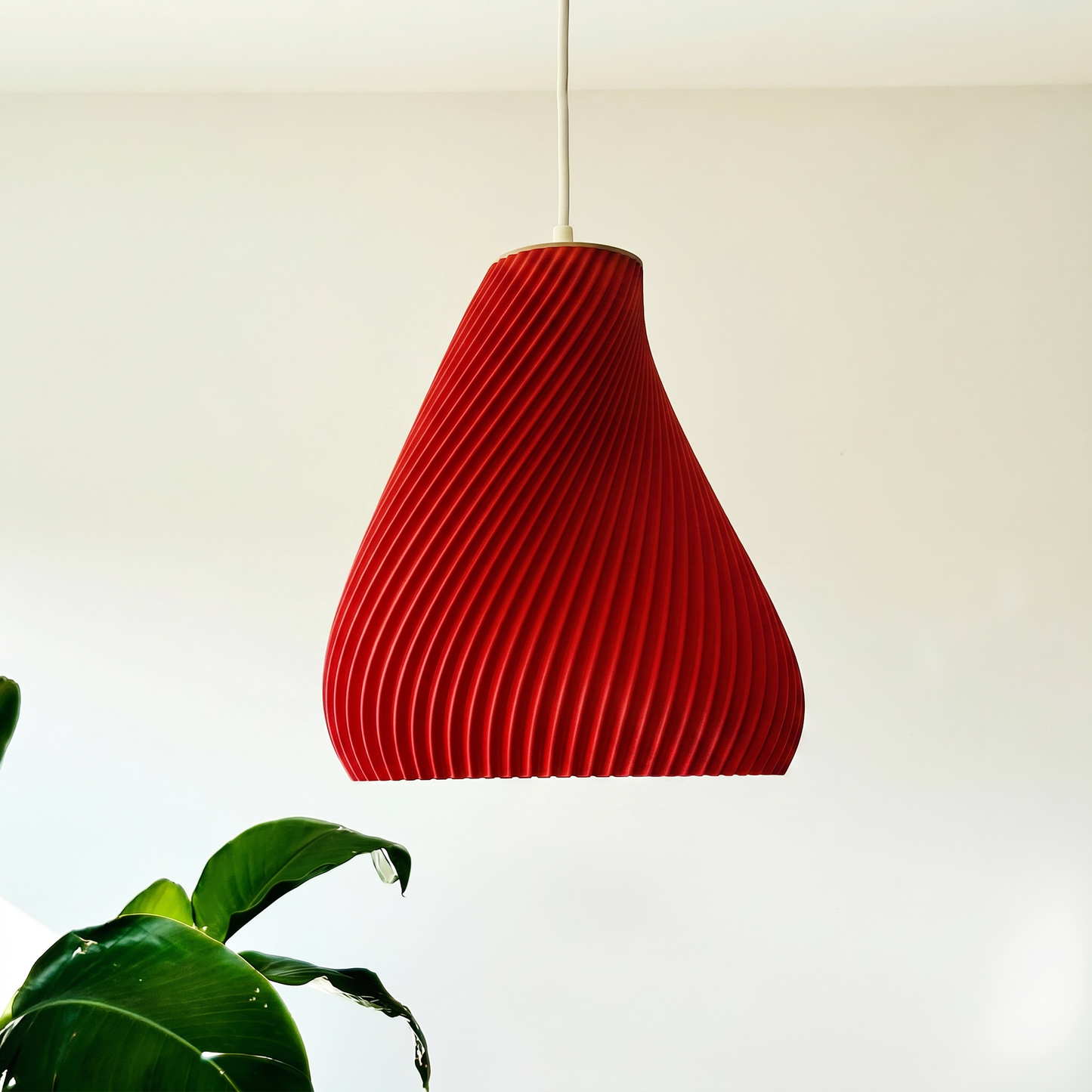 Red pendant light hanging against a plain background with a plant in the foreground.