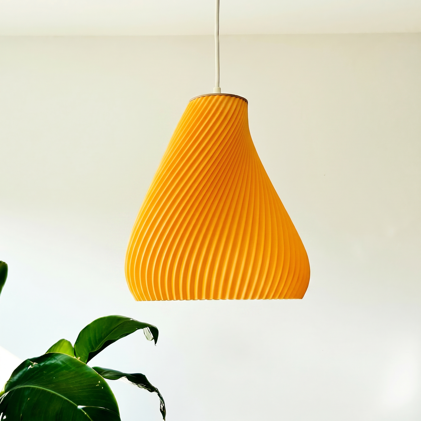 Orange pendant light hanging against a white wall with a plant in the foreground.